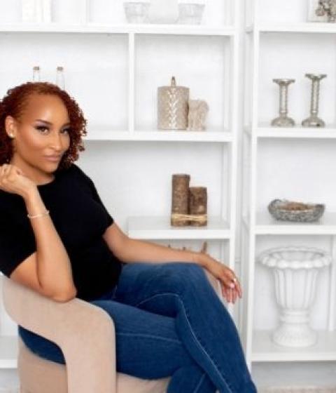 A person sits on a chair sideways in front of a shelf with books and decorative items.