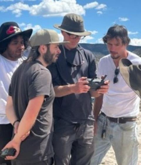 Four people standing in a huddle looking at a camera with a sweeping desert landscape behind them,