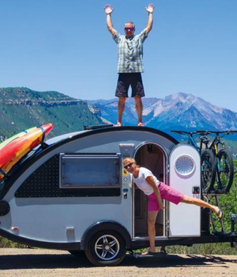 Earl stands atop camper with arms up and Glenna stands in door with leg out