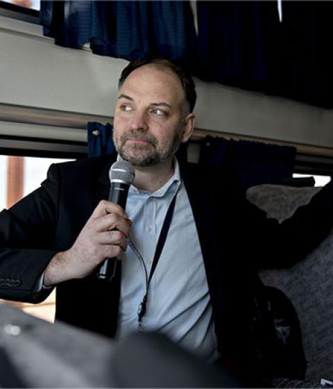 Stephen Gardner standing aboard an Amtrak train, holding a microphone in hand.