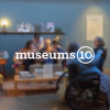 Museums 10 logo, white text with the word “museums” followed by a circle containing the number ten, overlaid on a blurred photo of museum visitors.
