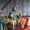 A person in a green “Sage” hoodie stands in a barn holding a cantaloupe and garlic, surrounded by farm gear on a red trailer.
