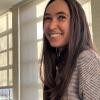 A person with long brown hair wearing a gray sweater smiles in front of a window.